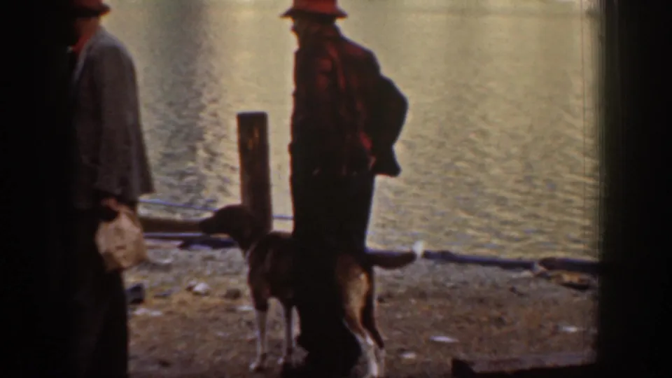 Two men in work jackets near a pier piling on a rocky shore, likely Lake Chelan, Washington, circa 1939; a dog stands in the foreground.