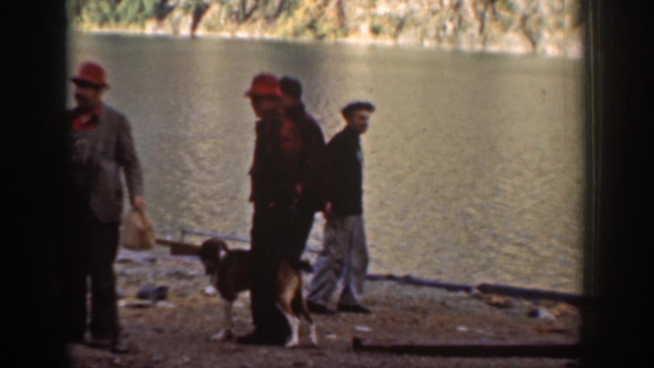Three men in brimmed hats on a rocky shore, a dog at their feet, with a calm lake and steep forested cliffs across the water.