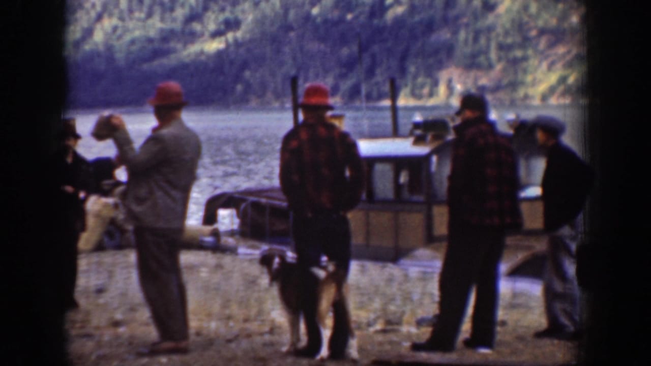 Four men in work clothing and brimmed hats on the rocky shore of what appears to be Lake Chelan, Washington, circa 1939; a utility boat is moored nearby and a dog stands among them.