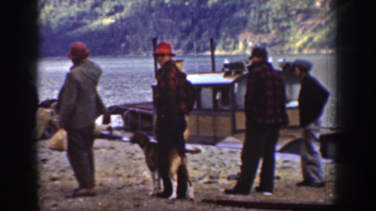 Four men in jackets and hats with a dog near a moored utility boat on what appears to be Lake Chelan, Washington, circa 1939; steep forested hills frame the background.