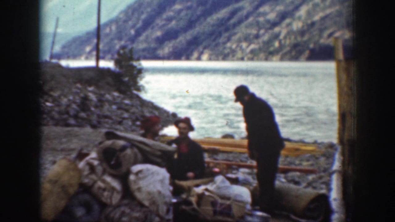 1939 color 8mm home movie: men resting on a rocky Washington lakeside with bundled gear, wooden poles, and forested mountains in the background.