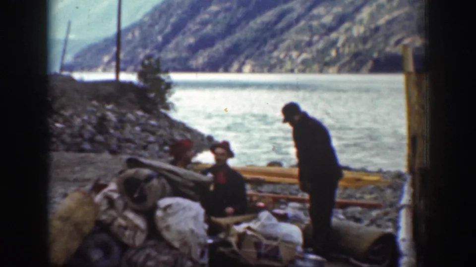1939 color 8mm home movie: men resting on a rocky Washington lakeside with bundled gear, wooden poles, and forested mountains in the background.