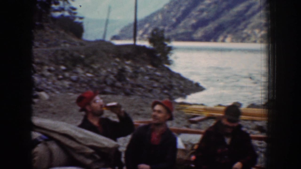 Two men seated close together, one raising a bottle to drink, both wearing red bandanas, lake and mountains behind.