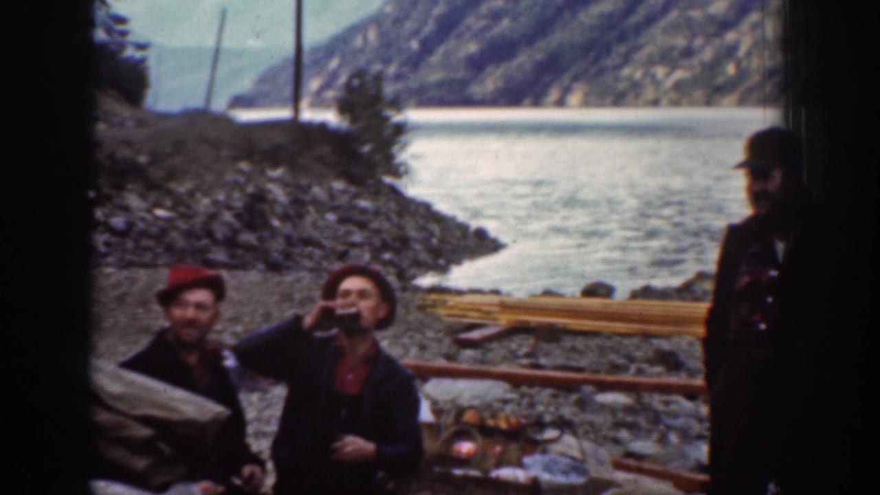 1939 8mm home movie: man in dark jacket drinks from a raised bottle on a rocky Washington lakeside, a second companion in hat visible at right, calm lake and forested hills behind.