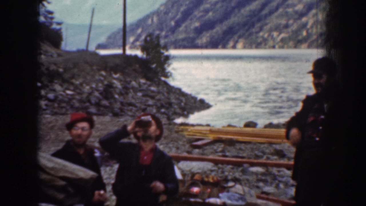 Three adult men on a rocky riverbank near Chelan, Washington, ca. 1939; two drink from bottles in matching red hats; river and forested mountains behind.