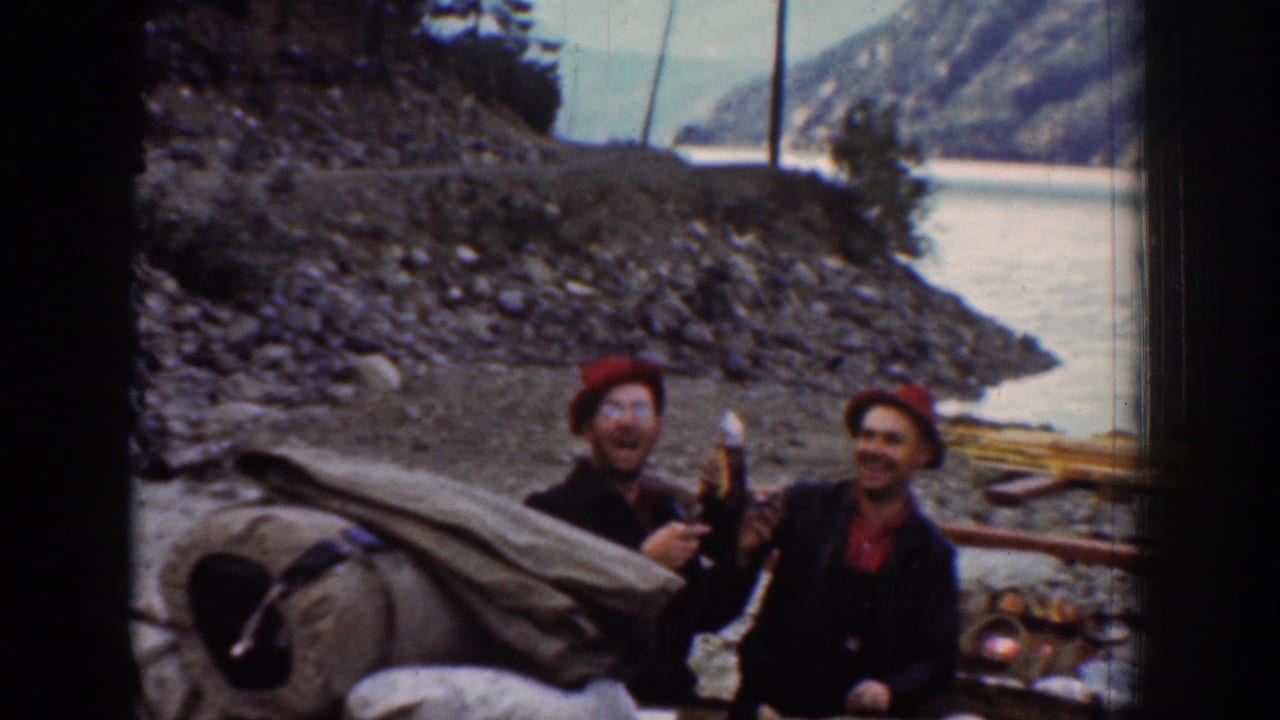 Two laughing men in red hats and dark jackets face the camera on a rocky riverbank; bundled canvas bedroll and camping gear visible behind them.