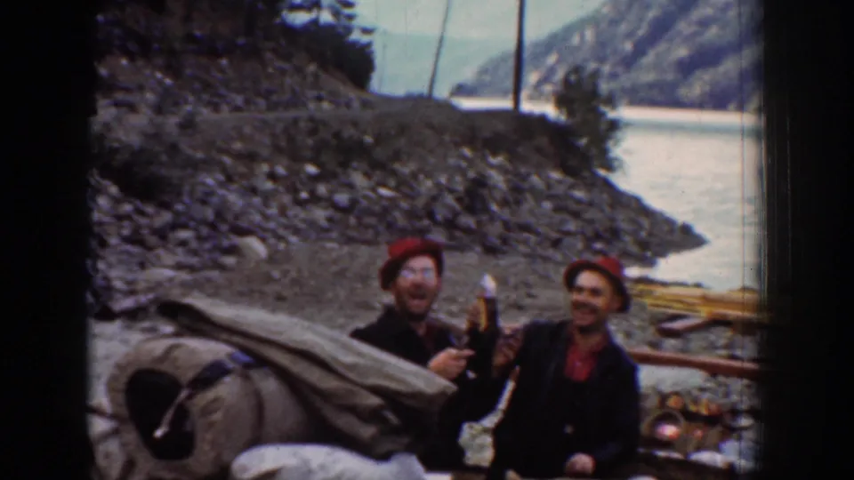 Two adult men, likely on a camping trip near Chelan, Washington, ca. 1939, laughing toward the camera; bundled canvas bedroll and river and mountains visible.