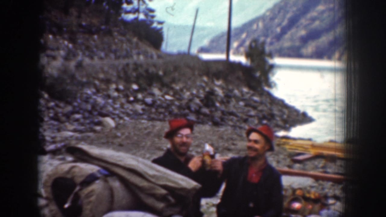 Two men on a rocky riverbank, possibly near Chelan, Washington, ca. 1939; one holds a bottle or snack; canvas bedrolls and river landscape visible behind.