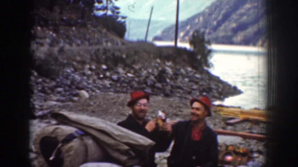 Two men on a rocky riverbank, possibly near Chelan, Washington, ca. 1939; one holds a bottle or snack; canvas bedrolls and river landscape visible behind.