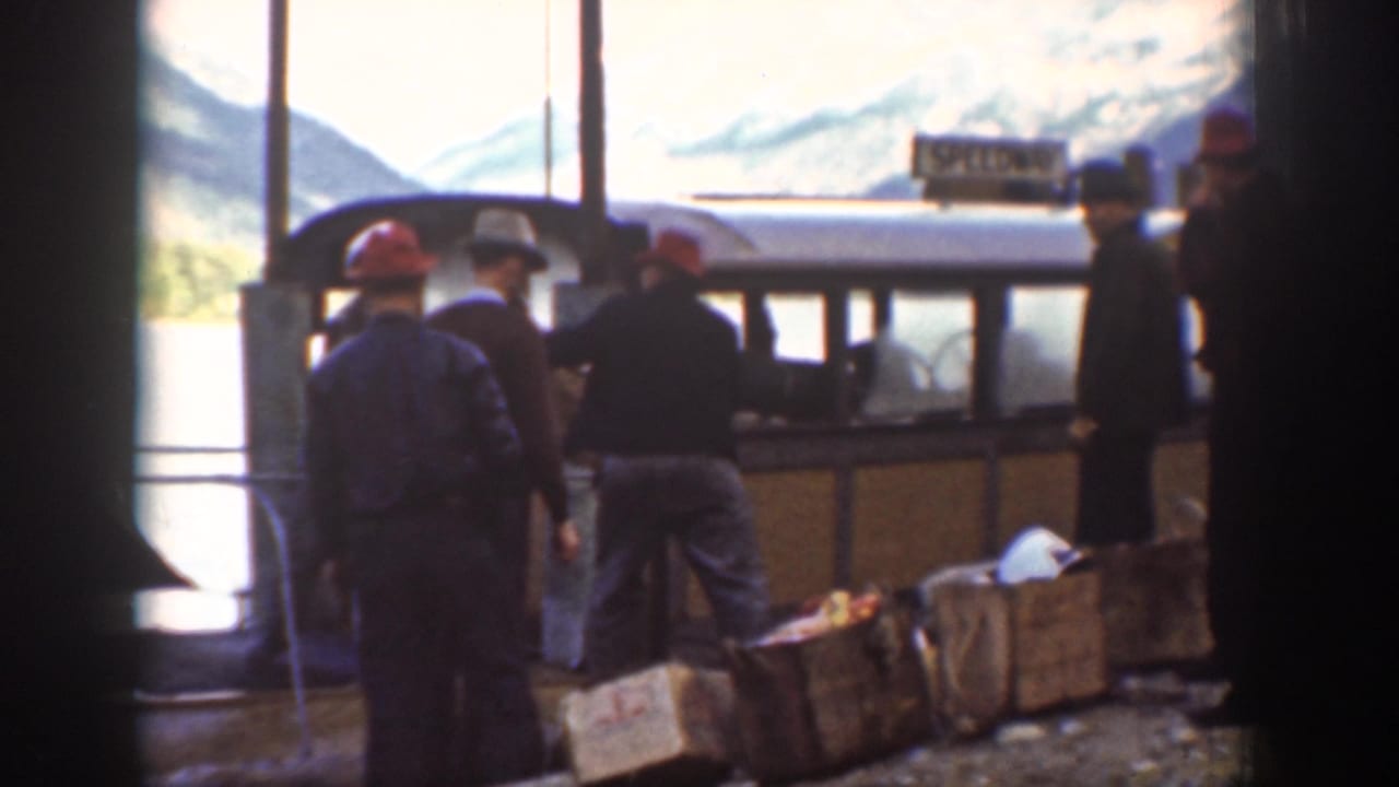 Group of men loading camping supplies near a roofed structure by the water, ca. 1939; wooden crates visible; mountains in background; partially legible sign requires review.