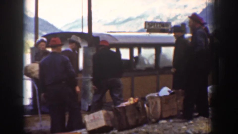 Workers load wooden crates alongside a touring bus at a mountain stop, Chelan, Washington, circa 1939; snow-capped peaks and a utility pole visible.