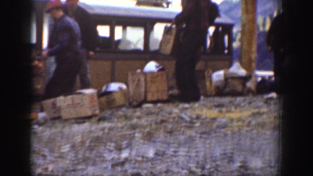 A worker in a red cap and dark coat stands near cargo crates at a mountain stop alongside a touring bus, circa 1939 Chelan, Washington.