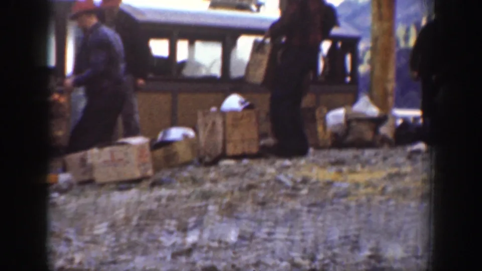 A worker in a red cap and dark coat stands near cargo crates at a mountain stop alongside a touring bus, circa 1939 Chelan, Washington.