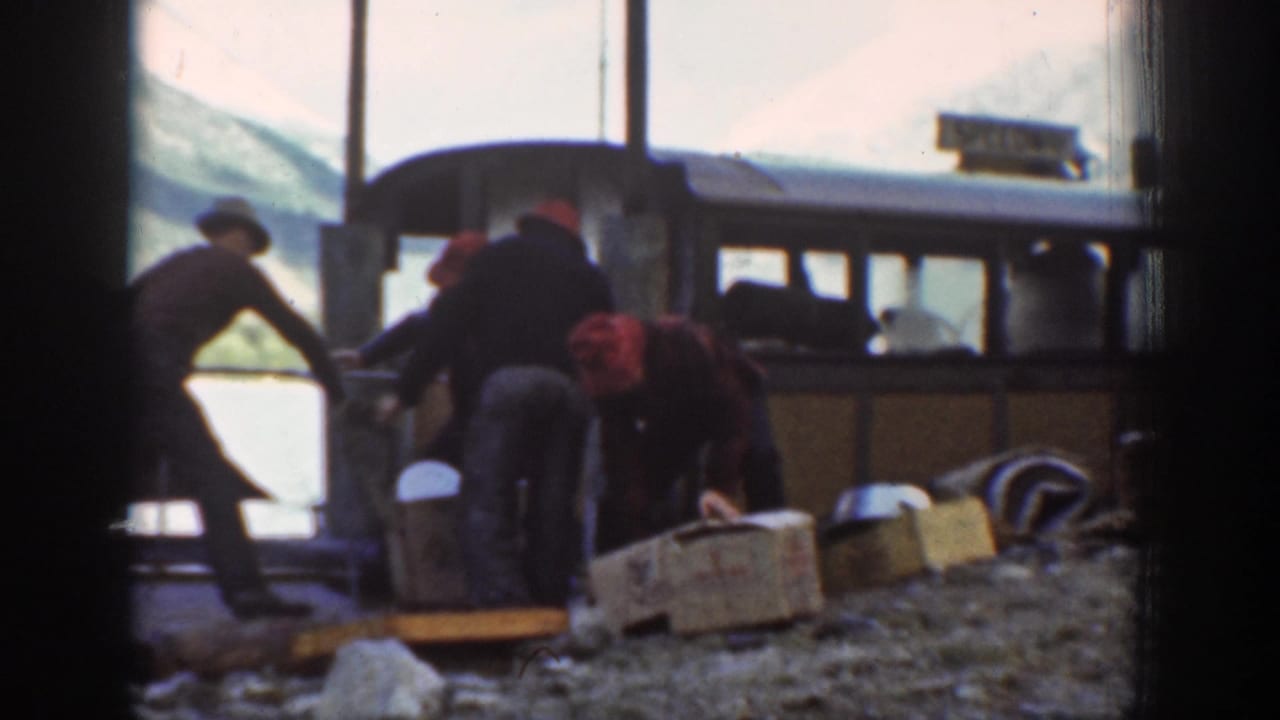 Men load wooden crates alongside a large windowed passenger vehicle; snow-capped peaks are visible above.