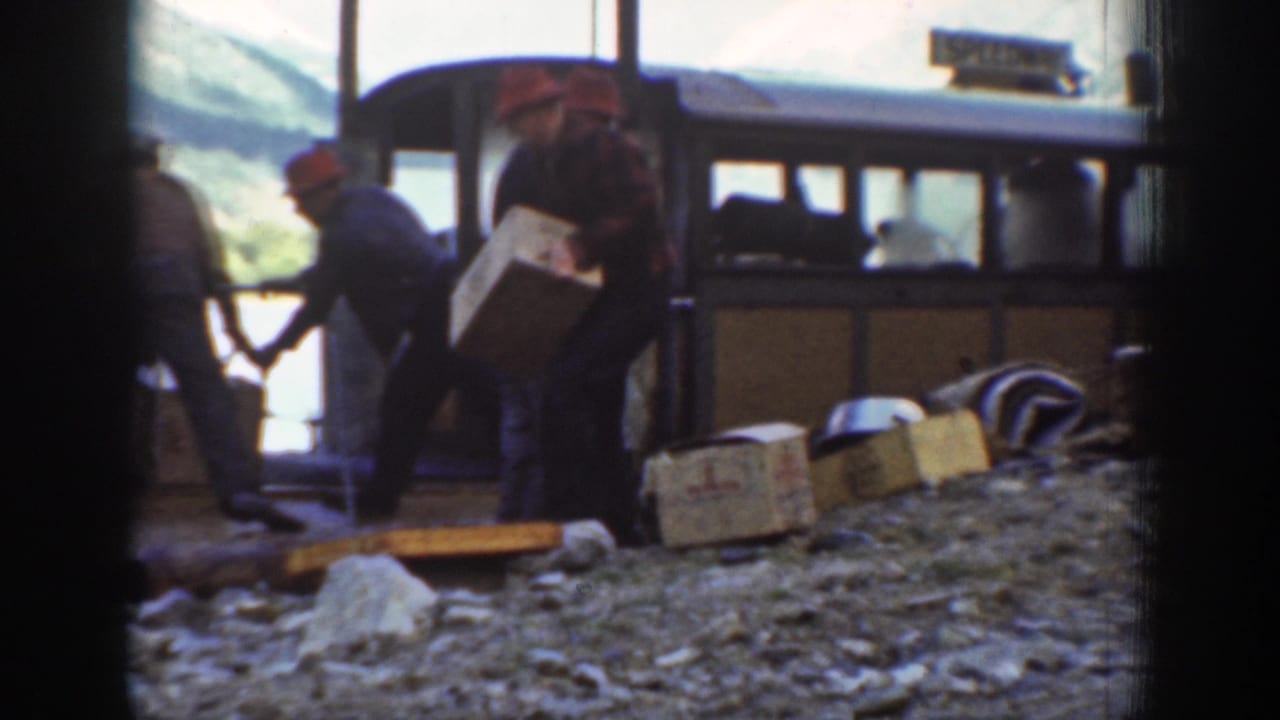 Workers with wooden crates alongside a 1930s-era passenger vehicle; a partially legible sign is mounted on the vehicle at a mountainous location.