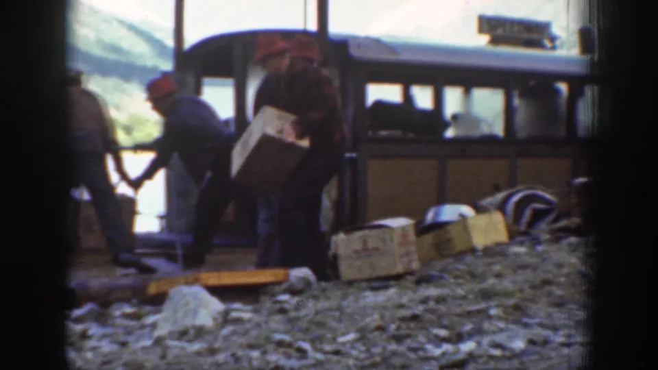 Workers with wooden crates alongside a 1930s-era passenger vehicle; a partially legible sign is mounted on the vehicle at a mountainous location.