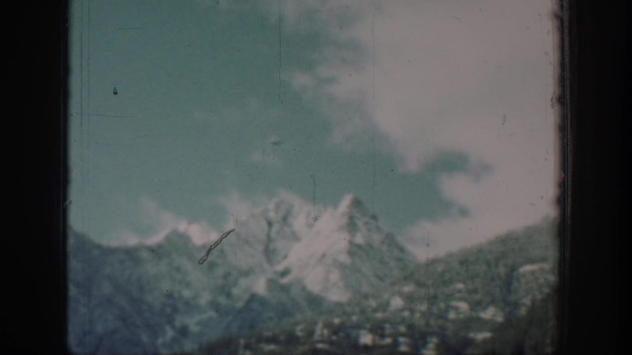 Passenger view from a boat on Lake Chelan, Washington, circa 1939, showing snow-capped peaks and forested slopes under a partly cloudy teal-blue sky, with a diagonal film scratch and 8mm grain.
