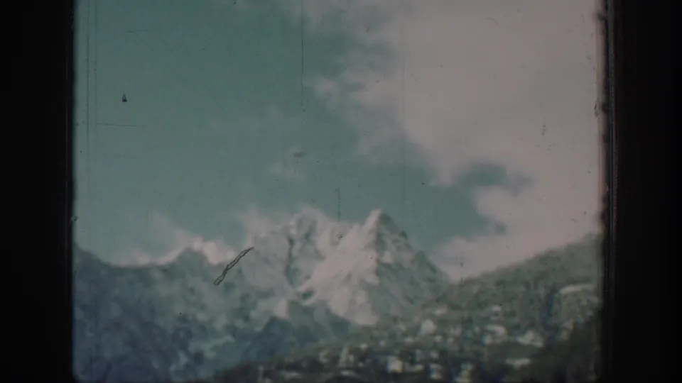 Passenger view from a boat on Lake Chelan, Washington, circa 1939, showing snow-capped peaks and forested slopes under a partly cloudy teal-blue sky, with a diagonal film scratch and 8mm grain.