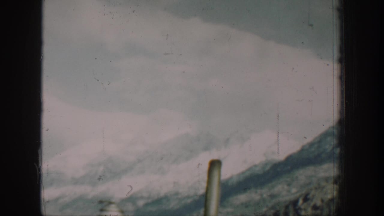 Overcast view from a boat on Lake Chelan, Washington, circa 1939, showing snow-covered mountain silhouettes and a single deck post, rendered in the muted tones of faded 8mm film.