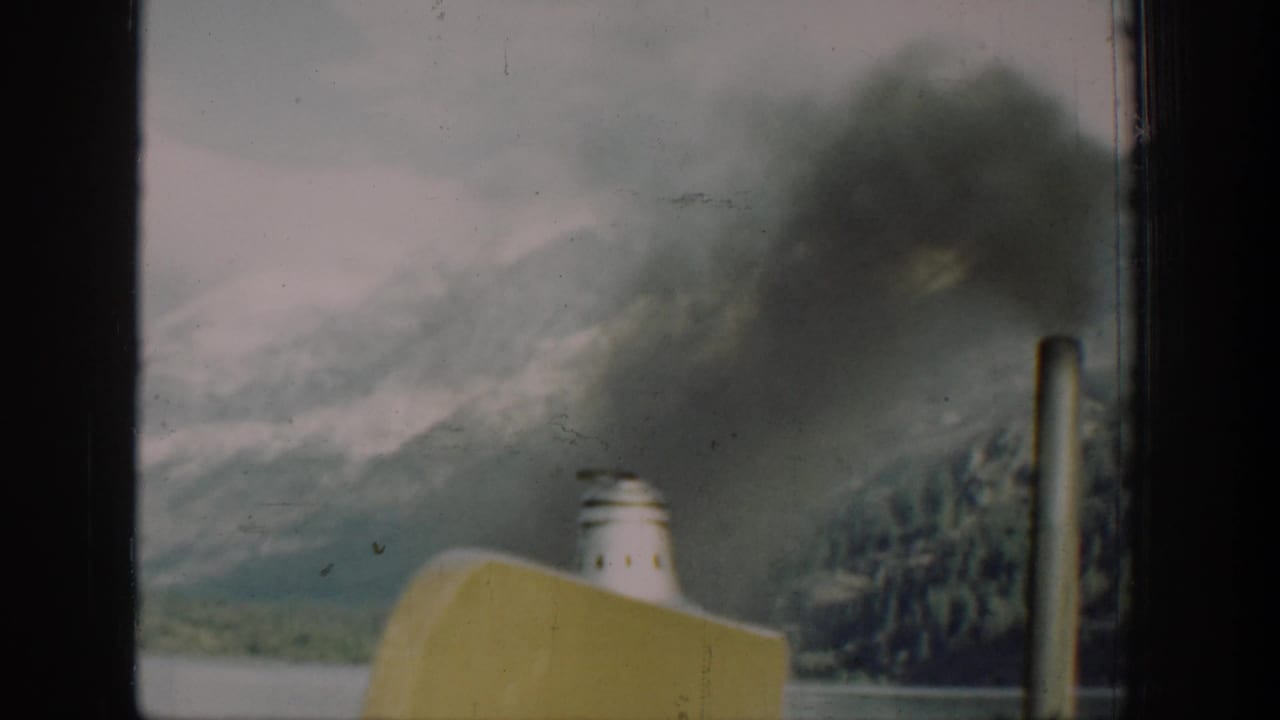 Boat deck detail on Lake Chelan, Washington, circa 1939: yellow hull, white smokestack emitting dark smoke, and mountain slopes visible beyond, captured on 8mm film with visible color fading.