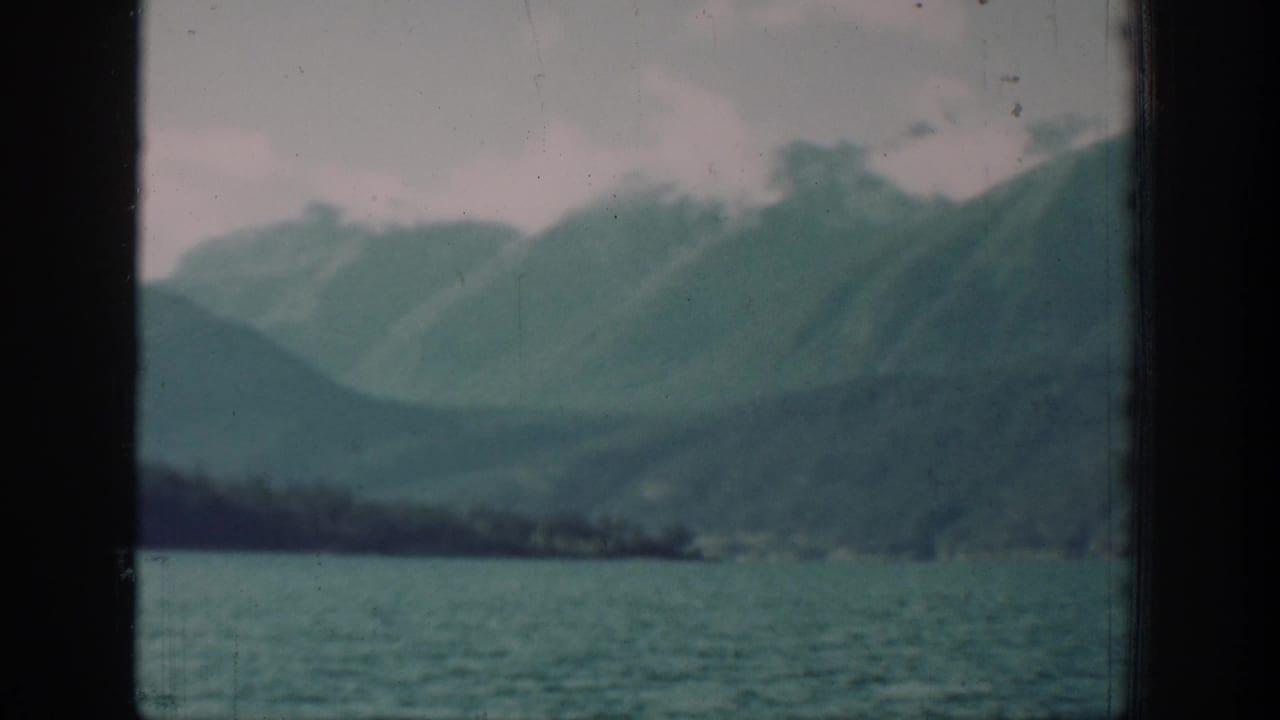1930s 8mm home movie wide shot of Lake Chelan area, Washington, showing a forested mountain ridgeline above calm lake water under a pale overcast sky.