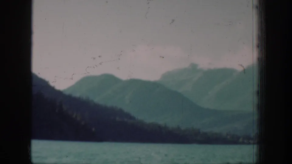 Archival 8mm home movie of the Lake Chelan area showing mountain slopes and lake with visible film scratches and a conifer treeline emerging at the right edge.