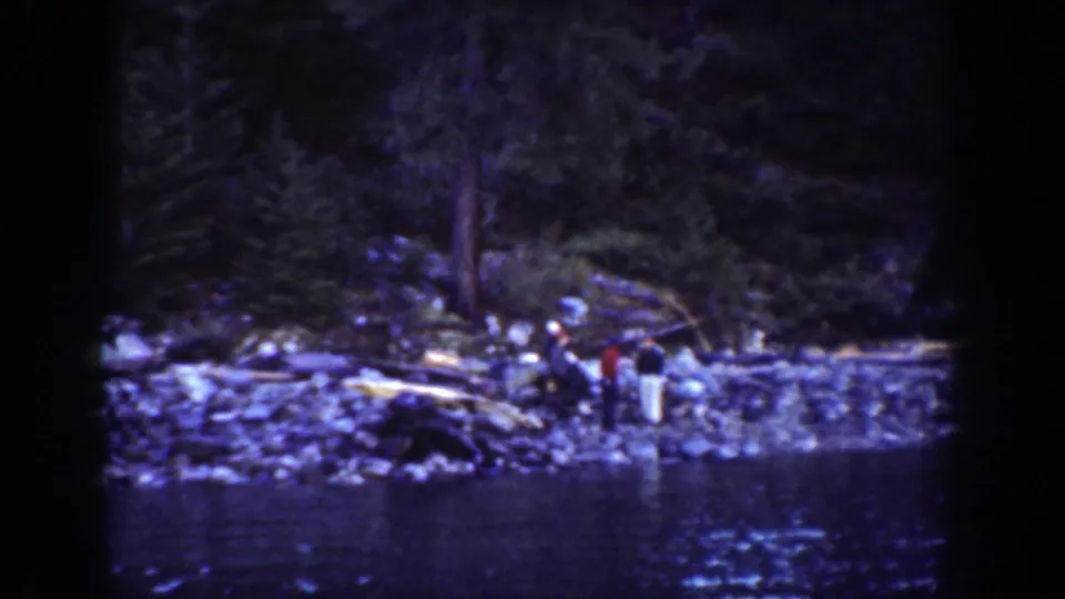 Archival 8mm home movie showing small figures on a rocky lakeshore near Lake Chelan, Washington, with dense conifer forest behind; faces not clearly discernible at this scale.