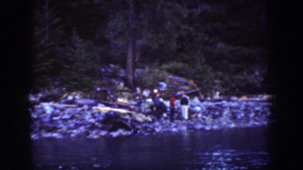Wide establishing shot of several figures on a rocky shoreline near Chelan, Washington, circa 1939; evergreen forest and lake water visible in background.