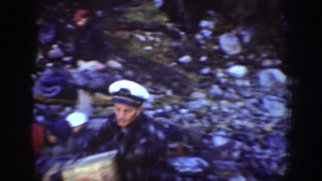 Man in white sailor cap and dark navy jacket carrying a wooden board on a rocky shore; second figure visible behind.