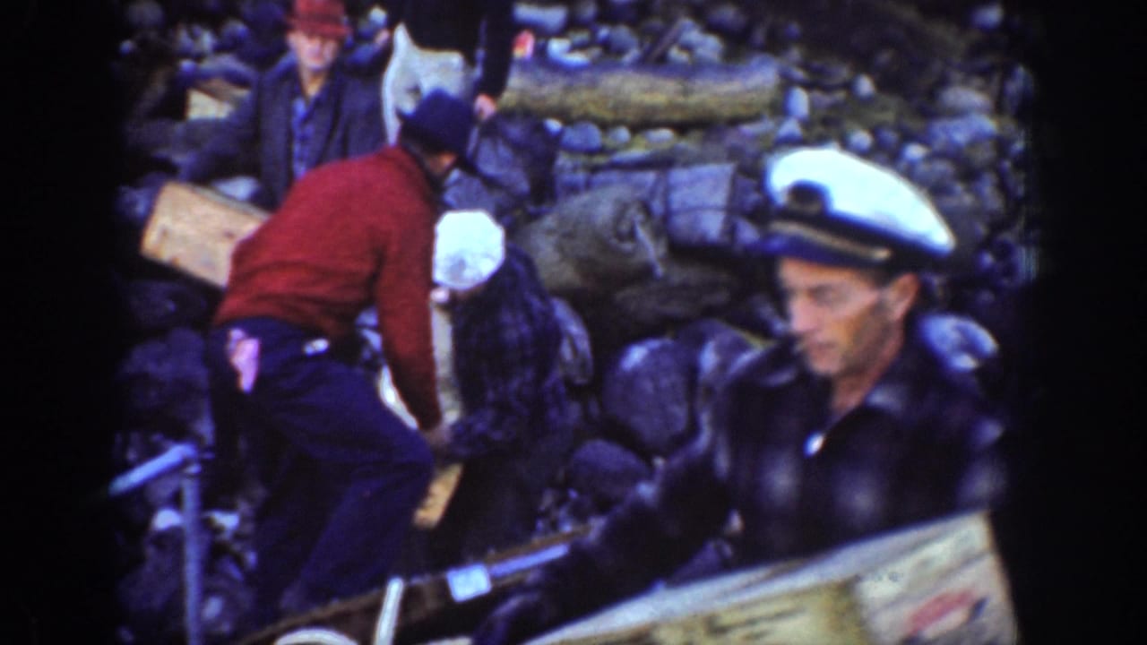 Group of men including a sailor in white cap and a companion in red sweater moving wooden crates on a rocky lakeshore, Chelan, Washington, circa 1939.