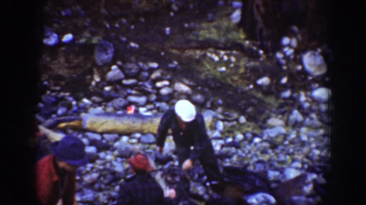 Sailor in white navy cap stooping on rocky lakeshore near Chelan, Washington, circa 1939; companion in dark jacket and fallen log visible beside him.