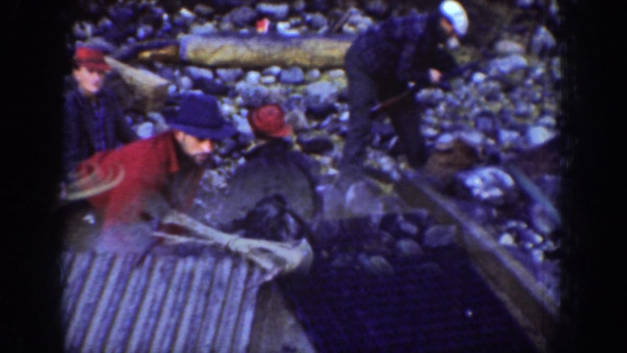 Three men in period work clothing handling corrugated metal sheeting in a rocky outdoor setting