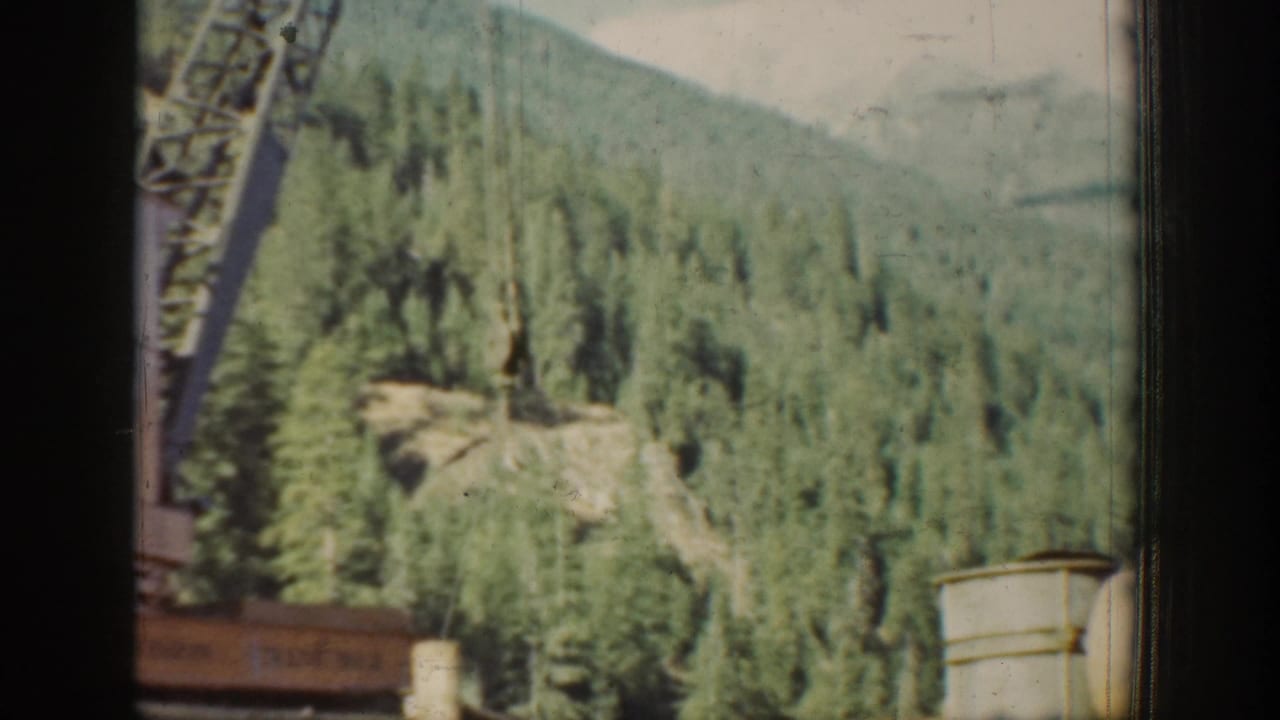 A tall lattice crane or derrick stands against a dense conifer-covered mountainside, consistent with a remote 1930s Pacific Northwest construction site