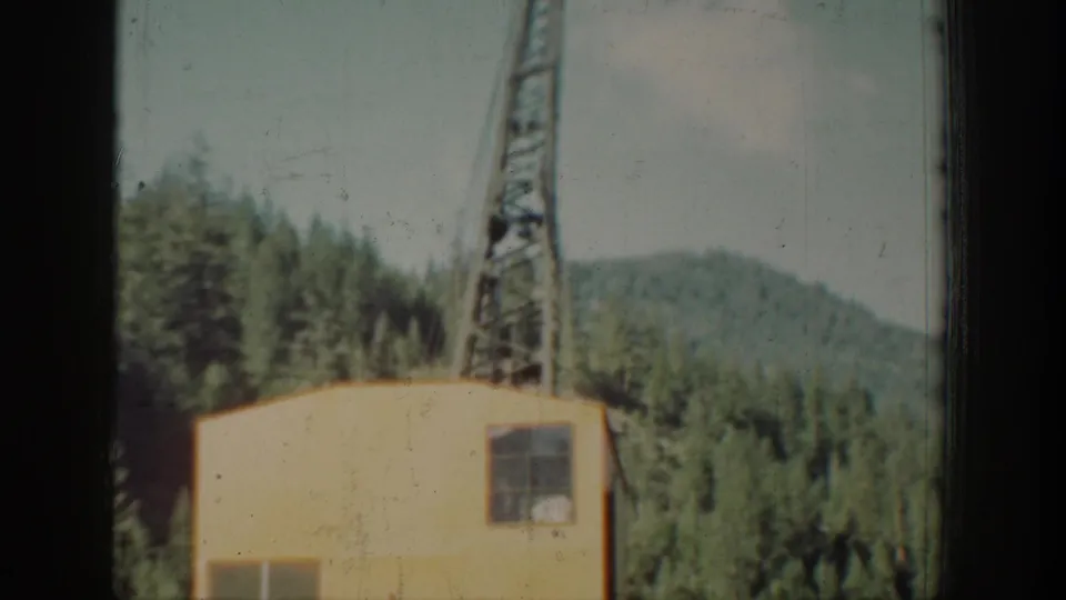 A lattice steel crane or derrick rises beside a small yellow outbuilding, set against a steep forested mountain ridge, ca. 1939, Washington state