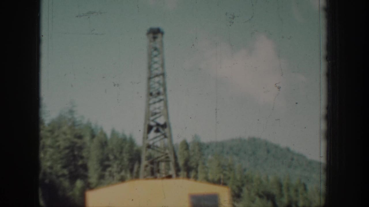 Yellow crane with tall steel lattice tower against a forested mountain backdrop, viewed through a window