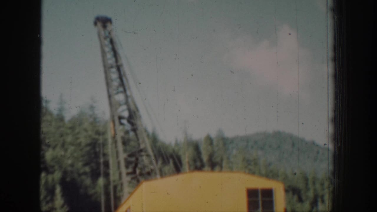 Yellow crane housing with angled steel lattice boom against conifer forest, shot through a window