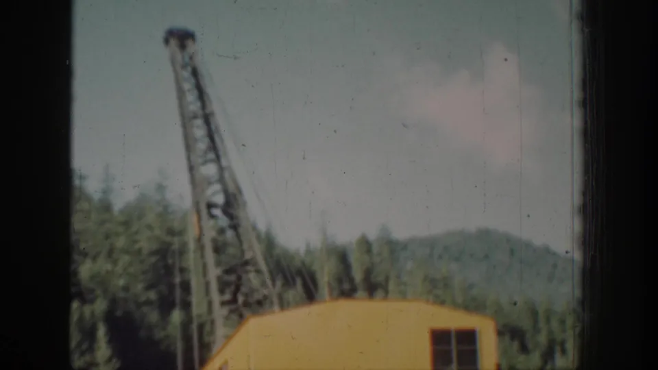 Yellow crane with lattice boom angled during load swing, Pacific Northwest conifer backdrop, Chelan area, circa 1939, 8mm color film