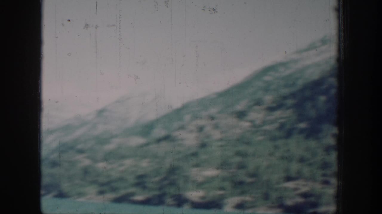 8mm color frame of green forested mountain slopes descending to calm lake water beneath a pale sky.