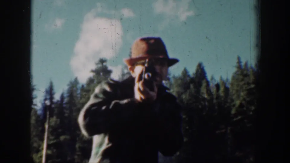 Adult male in wide-brimmed hat aiming a handheld camera toward the viewer, standing among Pacific Northwest evergreens under a blue sky with cumulus clouds, c.1939 8mm footage.