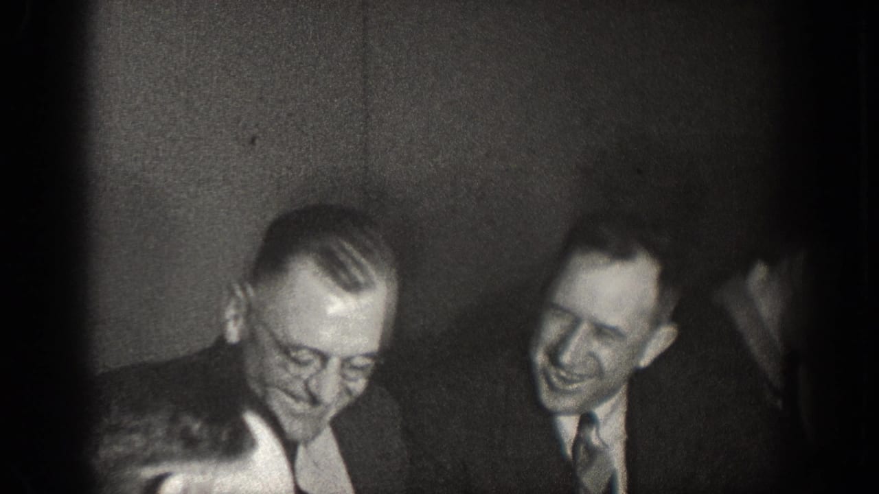 Black-and-white 8mm indoor meeting scene, circa 1930s — two men in lounge suits visible, one with eyeglasses, one smiling, at an unidentified civic or organizational gathering.