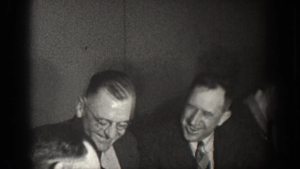 Black-and-white 8mm indoor meeting scene, circa 1930s — two men in lounge suits visible, one with eyeglasses, one smiling, at an unidentified civic or organizational gathering.