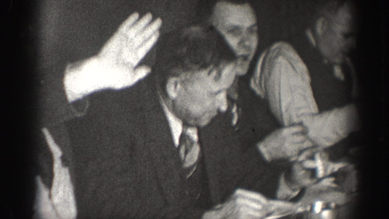 Black-and-white 8mm indoor meeting footage, circa 1930s — a man in a suit raises his hand while others at the table look on, suggesting a vote or procedural motion.