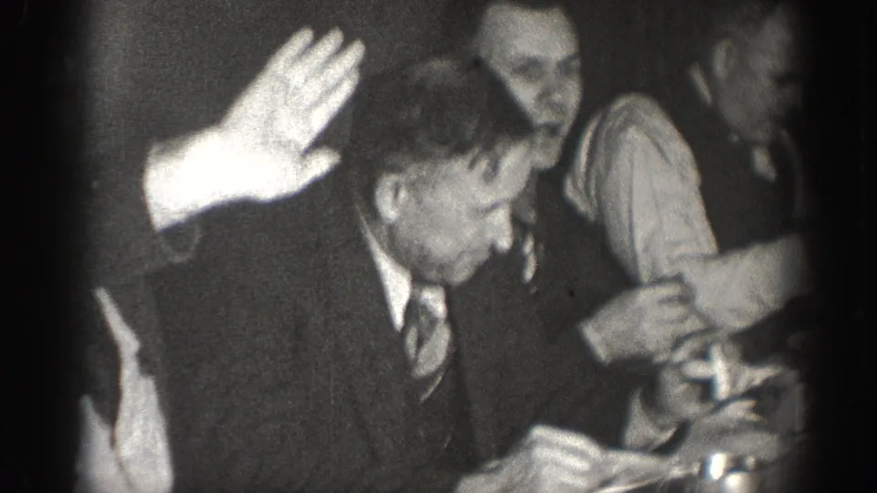 Black-and-white 8mm indoor meeting footage, circa 1930s — a man in a suit raises his hand while others at the table look on, suggesting a vote or procedural motion.