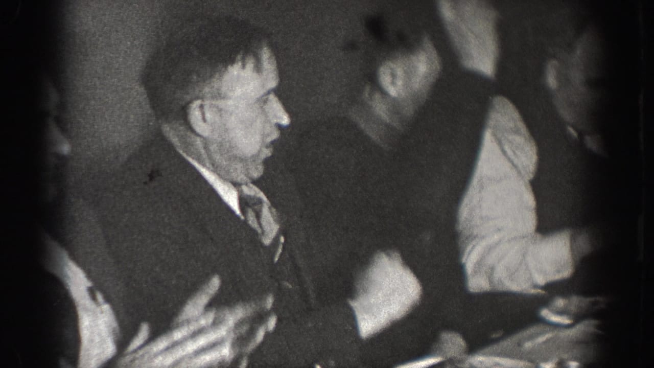 Several men at a dinner table; foreground figure in jacket and tie gesturing with both hands while conversing; heavy vignetting around frame edges.