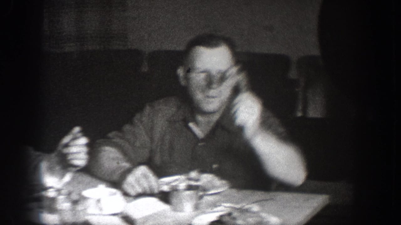 Man in glasses and dark shirt mid-gesture at a dinner table with dishes visible; circa 1939 home movie attributed to Chelan, Washington.