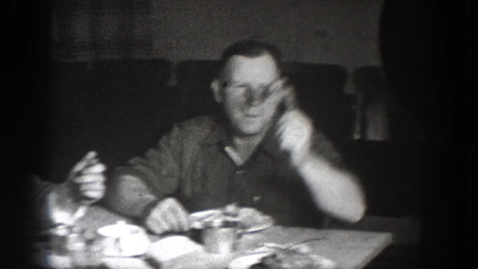 Man in glasses and dark shirt mid-gesture at a dinner table with dishes visible; circa 1939 home movie attributed to Chelan, Washington.