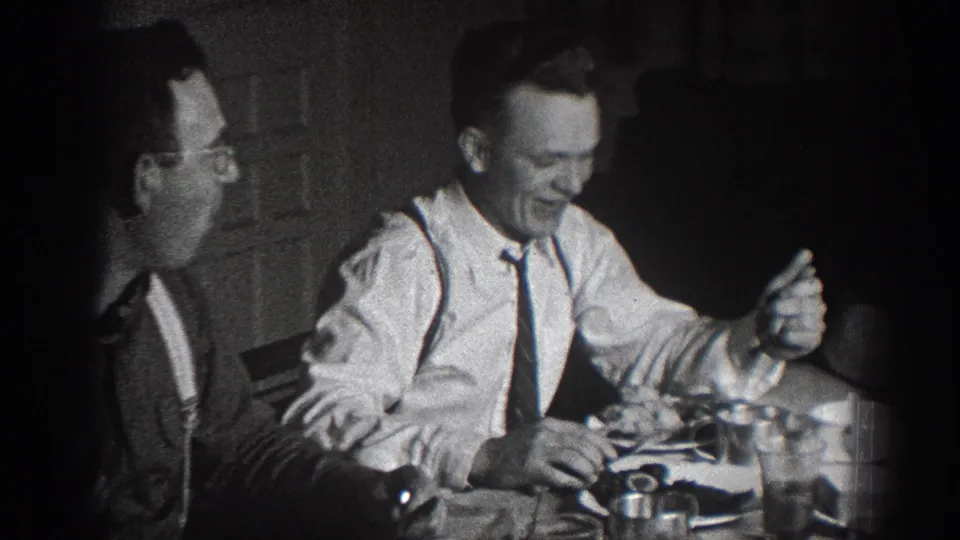 Two men in animated conversation at a dinner table, one in white dress shirt with suspenders and tie; late 1930s home movie attributed to Chelan, Washington.