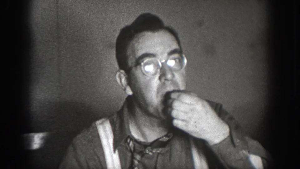 Man wearing round glasses, suspenders, and necktie eating or drinking at an indoor gathering, Chelan, Washington, circa 1939; black-and-white 8mm home movie.