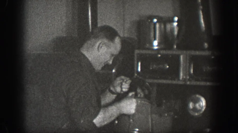 A man bends over what may be a stove or industrial appliance in a dimly lit interior, captured circa 1939 in Chelan, Washington, on 8mm film.