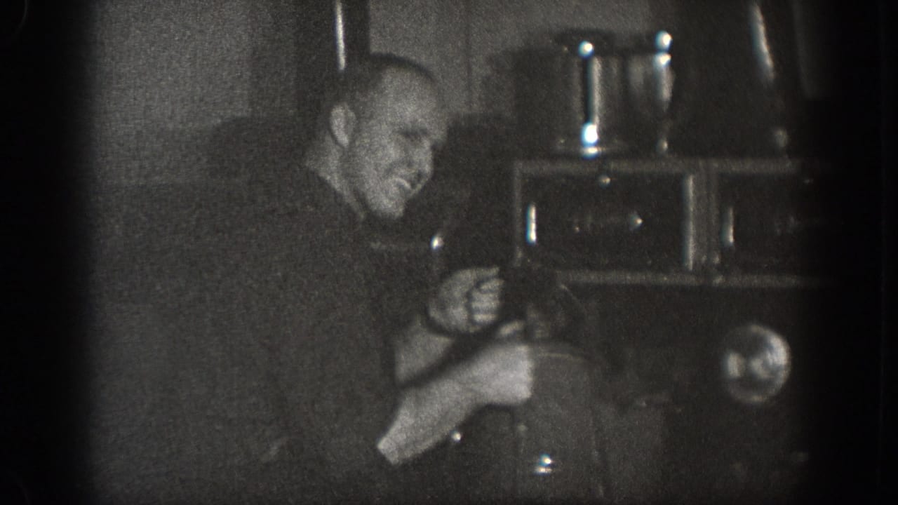 A man pauses to smile while working near a large dark appliance in a home movie from Chelan, Washington, dated 1939.